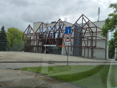 The image shows a street view in Šilutė, Lithuania, featuring the Šilutė Cultural Center. The building has a modern architectural design with a prominent brown, angular metal facade resembling abstract house frames. Large windows reflect the overcast sky. In front of the building, there is a paved area and a grassy verge. A traffic sign with a blue square and a white upward arrow is visible, along with a circular sign prohibiting cars and a sign indicating operating hours from 22:00 to 06:00. The text "KULTŪROS CENTRAS" and "CAFFEINE" are displayed on the building. The overall scene depicts a public space under a cloudy sky.