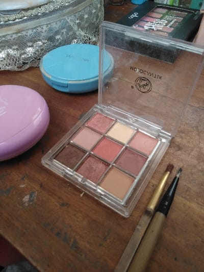The image displays a collection of cosmetic items arranged on a worn, dark wooden surface in Шөптікөл ауылдық округі, Kazakhstan. The central object is an open, clear plastic eyeshadow palette containing nine square pans of eyeshadow in shades including dark brown, shimmery copper, light pink, peach, cream, and medium brown. The transparent lid reflects the inverted text "WOLOCYMIK" and a circular logo featuring a stylized tree or plant design. To the right of the palette lie two makeup brushes with golden-brown handles and dark bristles, one appearing to be an eyeliner brush. To the left, a light pink oval-shaped compact is partially visible, and behind it, a light blue round compact with faint white text on its lid. In the upper left background, part of a silver-toned bag with a lace overlay pattern is visible. Further back, a black rectangular box, possibly another cosmetic palette, with the word "OSBO" printed on its side, is present. The wooden surface exhibits visible grain, scratches, and areas of wear and discoloration.
