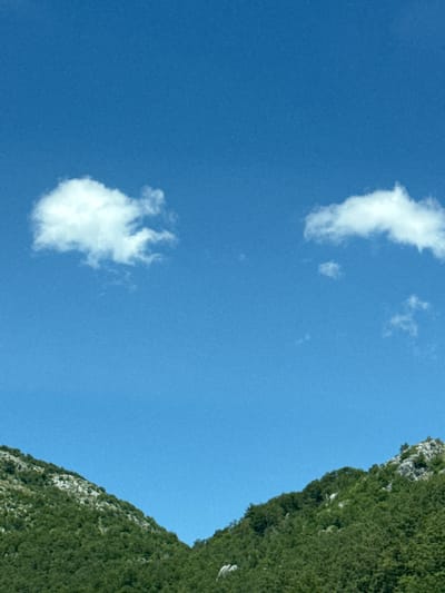 The image depicts a clear, vibrant blue sky with two scattered white cumulus clouds. The left cloud is larger and puffier, while the right cloud is smaller and more elongated. Below the sky, two densely forested mountain slopes converge towards the center, creating a V-shaped valley. The vegetation on the slopes appears lush and green, with some lighter-colored rocky outcrops visible on the upper parts of the mountains. The scene suggests a natural, outdoor environment, likely in a mountainous region. The location is specified as Obzovica, Montenegro. There are no people, animals, or man-made structures visible in the image.