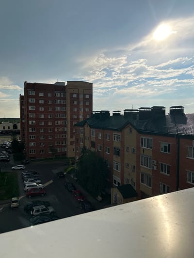 This is a medium-angle, outdoor, eye-level, vertical shot of apartment buildings in a residential area. The sun is bright in the sky, casting light and shadows. There are several buildings visible, some are red brick and others are yellow and cream colored. The buildings have many windows and balconies. Some of the buildings have dark green roofs. There is a street with cars parked on it and a tree. There are clouds in the sky. The scene appears to be taking place in the daytime. The text "ТОРГОВЫЙ ЦЕНТР" is visible on a building in the distance.