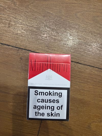 A Marlboro cigarettes pack is centered in the frame, resting on a wooden surface. The pack is predominantly red with a white diagonal stripe. The Marlboro logo is visible in red lettering across the top of the pack, and below the stripe, the words "SELECTED PREMIUM TOBACCOS" are printed in grey. A health warning, "SMOKING CAUSES AGEING OF THE SKIN," is displayed in black text on a white background at the bottom of the pack. The wooden surface exhibits a prominent grain pattern and appears to be a table or countertop. The lighting suggests an indoor setting. There are no people or specific landmarks indicating a location in Collado Villalba, Spain.