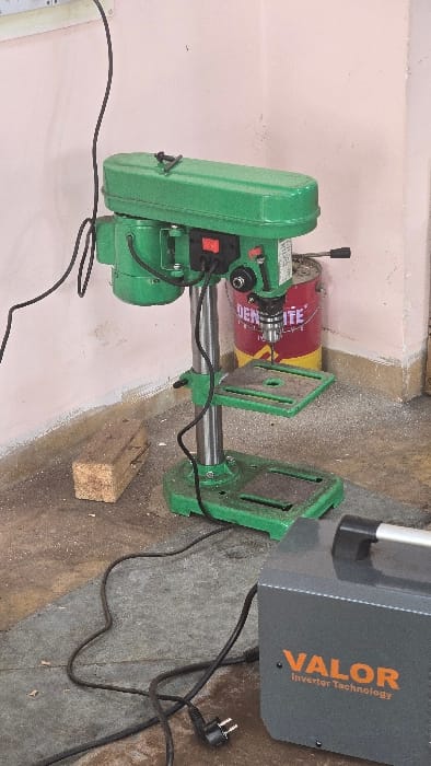 The image captures an indoor scene featuring various tools in what appears to be a workshop or utility area. A prominent green drill press is positioned on the left, displaying its chuck and a red power switch. Behind it, a red and yellow metal can has partially visible text including the word "LIFE."
To the right, a grey and orange machine is visible, clearly labeled "VALOR Inverter Technology," indicating it is likely an inverter welder or a similar piece of equipment. Black electrical cords are spread across the floor, with one terminating in a two-pin European-style plug. A small wooden block with nails embedded in it rests on the concrete floor.
The background consists of a light pink or peach-colored wall. The floor itself is a rough, possibly concrete surface, with some debris. No human activity is taking place; the tools are stationary. The scene suggests a functional workspace in an indeterminate time of day, consistent with an indoor setting, possibly located in Gangapur, India, as per the context.