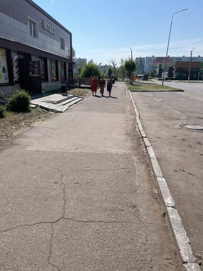 The image shows a street scene in Aksu, Kazakhstan, on a clear, sunny day. In the foreground, a cracked asphalt sidewalk runs along the right side of the frame, bordered by a low concrete curb. To the left, a paved pathway with visible cracks leads towards a building.
The building on the left is a commercial establishment with a sign reading "OCCBET". It features large display windows showcasing various products. In front of the building, a small green bush is visible, and a white cat is sitting on the stone steps leading to the entrance.
Three women are walking on the pathway towards the right side of the image. The woman on the left is wearing a red dress, the middle woman is in a brown outfit, and the woman on the right is wearing a floral dress and a patterned headscarf.
Further down the street, on the right side, there is a road with some parked cars and modern buildings in the background. Power lines are visible crisscrossing the sky. In the distance, apartment buildings with colorful facades and a large mural depicting a person's face are notable features of the urban landscape. The environment appears to be a typical urban street with commercial and residential elements.