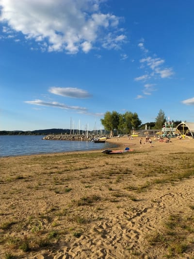 The image depicts a sandy beach on a sunny day, with a calm body of water to the left and a clear blue sky with scattered clouds above. In the background, a rocky breakwater separates the beach from a body of water where several sailboats are moored. Trees and hills form the distant shoreline.
On the beach, a small group of people are engaged in various activities. Several individuals are lying on beach towels, relaxing. Some children are playing in the sand, one appears to be kicking a ball, while others are seated or standing. There is a structure that resembles a lifeguard tower or a small observation deck with a yellow slide. Further to the right, a large tent-like structure with a thatched roof is partially visible. A few surfboards or paddleboards are lying on the sand. The overall scene suggests a recreational area, possibly a beach resort or a public park with a lake. The mention of Radslav, Czechia, places this scene in that specific geographical location, implying it is a popular spot for locals and tourists to enjoy outdoor activities during warmer weather.