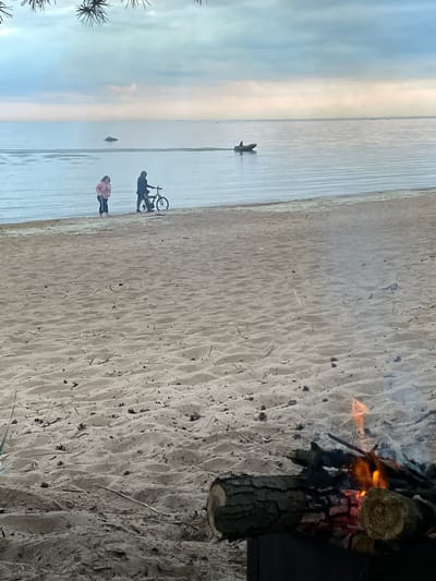 The image depicts a sandy beach with a calm body of water in the background. Two individuals, one wearing a pink hoodie and the other in dark clothing, are standing near the water's edge, with one of them beside a bicycle. Further out on the water, a small boat with a person inside is visible. In the foreground, a campfire is burning, with logs and branches arranged for fuel. The sky is overcast with a soft, diffused light. The location is identified as Chernaya Lakhta, Russia.