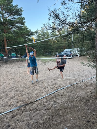 In Ozerki (Krasnodolinskaya), Russia, four individuals are engaged in a game of beach volleyball on a sandy court surrounded by pine trees. A volleyball is suspended in the air above the net, indicating an active play. Two men are on one side of the net, one in a blue tank top and shorts appears to be mid-jump, reaching for the ball, while another man in a grey t-shirt and black shorts is kicking the ball. On the other side of the net, a woman in a pink t-shirt and dark pants and a man in a light-colored t-shirt and blue shorts are positioned on the court. In the background, tents and a car suggest a campground or recreational area. The sky is clear and blue.