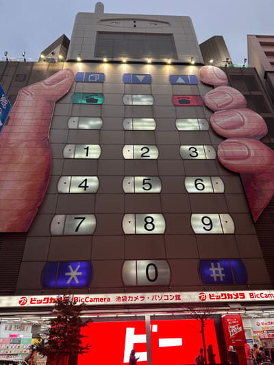 This image shows the facade of a building designed to resemble an oversized mobile phone. A giant hand graphic is present, with fingers depicted as pressing the numerical keypad. The buttons are arranged in a typical phone layout, with numbers 1 to 0, plus asterisk and hash symbols. Adjacent to the building is signage for Bic Camera, indicating the store's location in Ikebukuro, and advertising its camera and computer products.