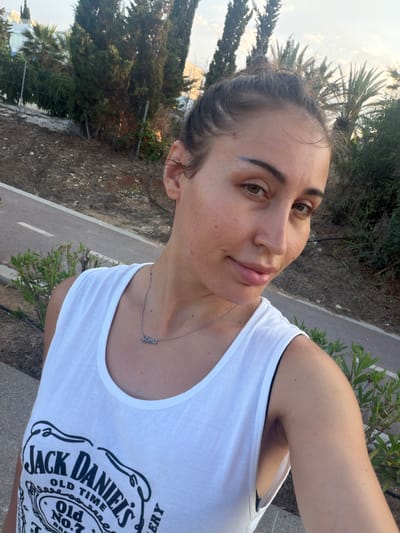 A woman with her hair pulled up in a bun takes a selfie outdoors in Journalists Village, Cyprus. She is wearing a white tank top with a graphic print of the Jack Daniel's Old No. 7 Tennessee Whiskey logo. Around her neck, she wears a silver necklace with the word "Mama" in cursive. The background consists of trees, a paved path, and a chain-link fence. The lighting suggests it is either early morning or late afternoon.