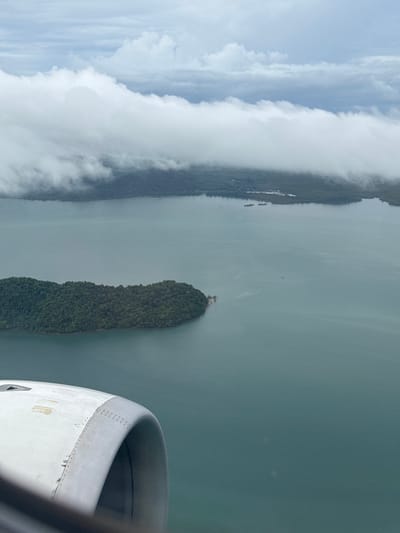 This is an aerial view from an airplane window, looking out over a body of water and a densely forested island. The water is a muted, greenish-blue, with some slight ripples visible. A long, narrow island covered in lush green trees dominates the center of the frame, its shape elongated and somewhat pointed at one end. Atop this island, a small cluster of structures, possibly buildings or a small settlement, is visible. In the distance, the coastline of what appears to be a larger landmass is partially obscured by thick, white clouds that hang low and stretch across the frame, obscuring the horizon. The sky above the clouds is a pale blue, suggesting it is daytime. The edge of an airplane engine cowling is visible in the lower right corner of the image, indicating the perspective is from inside a moving aircraft. The overall scene conveys a sense of remote, tropical natural beauty, with a hint of civilization present on the island. The weather appears to be partly cloudy, with the clouds creating a dramatic contrast with the water and the island. No text is visible in the image.