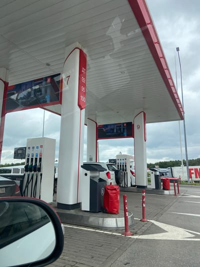 The image shows a gas station in the city of Uzytamak, Russia. The gas station has red and white columns with numbers indicating fuel types. There are several cars at the pumps, and a truck in the background. The sky is cloudy. The scene is orderly and functional, depicting a typical gas station environment.