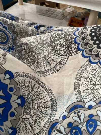 The image presents a close-up, angled view of a folded textile fabric, located in Sredkovets, Bulgaria. The fabric features a repeating pattern of intricate, mandala-style circular and semi-circular motifs. The primary colors in the pattern are various shades of blue (including royal blue and lighter blues), grey (dark and light), and black for outlining and finer details, all set against an off-white or light cream background. Visible creases and folds indicate the fabric is draped or resting on a surface. In the heavily blurred background, a light-colored surface, consistent with a workbench or table, is discernible. On this surface, indistinct objects are present, including a white spool of thread positioned to the left, flat items with green markings, and several small, colorful objects further back, suggesting a workspace or crafting environment. No individuals, explicit actions, or direct interactions are visible.