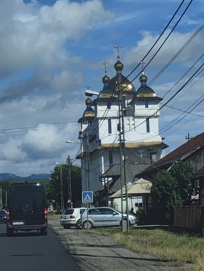 The image captures a street scene in Sighetu Marmatiei, Romania, featuring a prominent white church with multiple golden domes topped with crosses, against a backdrop of a blue sky with scattered clouds. In the foreground, a black Mercedes-Benz Sprinter van is parked on the left side of the asphalt road, displaying a license plate that reads "FF 491U". To the right of the van, a white Volkswagen Golf and a silver car are parked on the side of the road. A pedestrian crossing sign is visible next to the parked cars. The church structure is multi-tiered, with several windows visible on its facade. Overhead, a network of power lines crisscrosses the sky, partially obscuring the church and the sky. Adjacent to the church, there are other buildings, including a house with a brown tiled roof and a structure with a light-colored awning. Green trees and vegetation are scattered throughout the scene. In the distance, hazy mountains can be seen under the cloudy sky. The overall impression is a typical roadside view in a town or city, with religious architecture, vehicles, and everyday infrastructure coexisting.