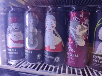 The image shows a close-up of several cans of craft beer sitting on a white, wire rack inside a refrigerated display. The cans appear to be in a shop in Pskov, Russia. The visible brands include “БAБA ЯГА Breakfast Stout,” “MИКИМОРА Stout," "MИЛИСA Oatmeal Stout,” “PУСАЛКА Sour Ale,” and “BEHEBИ Black IPA”, all brewed by BREWLOK. Each can features a unique stylized illustration of Slavic folklore figures. The scene is brightly lit from within the display case.