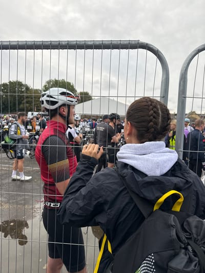 The scene depicts an outdoor sporting event, presumably a triathlon, located in Weymouth, United Kingdom, under an overcast sky with puddles visible on the paved ground. A galvanized metal crowd control barrier occupies the foreground, separating observers from the event area.
In the right foreground, an individual is seen from behind, featuring dark hair styled in two French braids, wearing a light grey hooded sweatshirt layered under a black waterproof jacket. A black backpack with yellow straps and a white dotted design is on their back, and their right hand rests on the top rail of the barrier. To the left foreground, a male in profile wears a white cycling helmet with "TEAM BYTE" branding, a red and black cycling jersey with horizontal yellow stripes on the sleeves, and black cycling shorts with "ELITE3" on the waistband. He has a dark beard. Both individuals are positioned at the barrier, observing the activity beyond it.
Beyond the barrier, a crowded area contains numerous participants and officials. Individuals wear varied sportswear, including one person in a black shirt labeled "TECHNICAL OFFICIAL" and another with "RUN" on their shorts. Many bicycles are present, indicating a transition zone or event staging area. White event tents are visible in the mid-background, with green trees and light-colored buildings further in the distance. The ground beyond the barrier is also paved and appears damp.