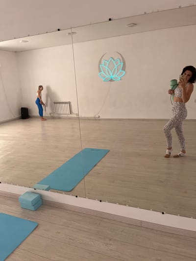 In a brightly lit, spacious room with light wooden floors and white walls, a woman in a leopard-print sports bra and matching leggings stands facing a large mirror, holding a green yoga mat and a phone. She is taking a selfie. Reflected in the mirror, a second woman in a white sports bra and blue leggings stands with her back to the mirror, holding a water bottle. A blue yoga mat is laid out on the floor in front of the mirror, and a blue yoga block sits beside it. A neon lotus flower light is mounted on the wall. A black speaker and a white radiator are visible in the background. The room appears to be a yoga or fitness studio. This image was taken in Aksu, Kazakhstan.