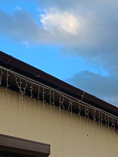 The image shows a section of a light-colored building exterior with a dark brown eave and gutter. A string of white icicle lights is attached along the underside of the eave. The background is a blue sky with scattered clouds. The location is identified as Zelenchukskaya, Russia.