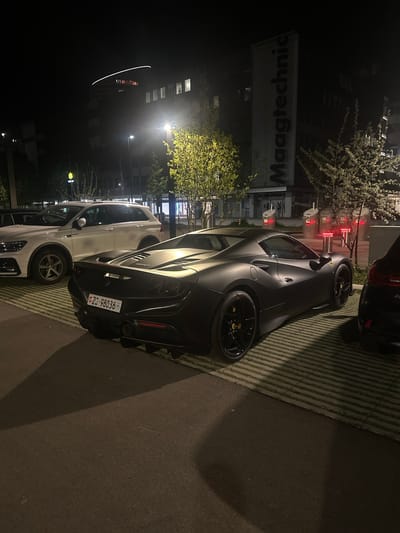 The image depicts a nighttime outdoor scene, likely a parking area or street, in an urban environment. The primary subject is a sleek, matte grey sports car, identified by a yellow emblem on its black multi-spoke wheels, strongly suggesting a Ferrari. The car is parked on a patterned surface, possibly grass pavers, and displays a Swiss license plate "ZG - 98036" on its rear. Yellow brake calipers are visible behind the spokes of the wheels.
Behind the sports car, a white SUV is parked, its front partially obscured. A dark-colored vehicle is also partially visible in the upper right background. The scene is illuminated by artificial light sources, including streetlights, creating good visibility despite the dark sky.
In the background, several multi-story buildings stand, with some windows aglow. A prominent sign on one building clearly displays the text "Maagtechnic." Trees with sparse or newly budding leaves are scattered around, catching the light. There is no visible human activity or specific event taking place; the vehicles appear to be stationary and parked. The atmosphere is calm and quiet, characteristic of a late-night setting.