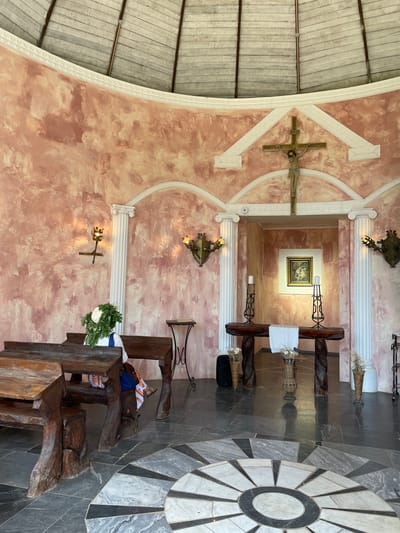 The image depicts the interior of a chapel with a rounded, textured pink wall. In the center of the back wall, a wooden crucifix with Jesus is mounted. Below the crucifix, an arched doorway reveals a smaller room with a framed painting of two figures. In front of this doorway is a wooden altar with two tall candle holders and white cloths. Flanking the altar are two white columns. Two wall sconces with flame-like lights are present on the side walls. In the foreground, two wooden benches with carved legs are positioned, with a figure partially visible sitting on one of the benches, adorned with a green leafy headdress. The floor is tiled with a geometric pattern. The ceiling appears to be made of wooden planks. The overall ambiance suggests a place of worship, possibly a small chapel or an oratory.