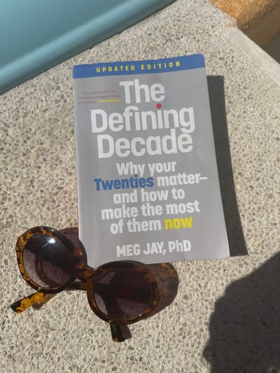 The image depicts a book titled "The Defining Decade: Why your Twenties matter--and how to make the most of them now," written by Meg Jay, PhD. The book is an updated edition and is described as "A four-alarm call for the 50 million twentysomethings in America" by Kirkus Reviews. A pair of round, tortoiseshell sunglasses rests on top of the book. The setting appears to be outdoors, likely on a sunny day, as indicated by the presence of shadows.