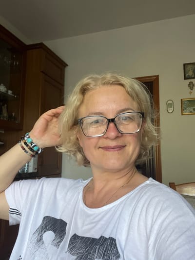 A close-up, waist-up, selfie-style photograph shows a woman with blonde, curly hair and glasses. She is wearing a white t-shirt with a black graphic design and a gold necklace. On her left wrist, she is wearing multiple beaded bracelets. Her left arm is raised and her hand is touching her hair. She is smiling and looking at the camera. The background appears to be indoors, with wooden cabinets and framed pictures on the wall, suggesting a domestic setting. The lighting is natural and coming from the right side of the frame. The image captures a candid moment of the woman.