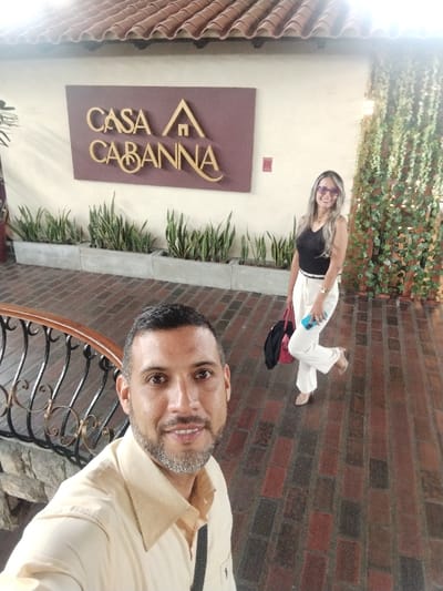A selfie shows a man with short dark hair and a light tan shirt in the foreground. Behind him, a woman in a black top and white pants poses near the "Casa Cabanna" sign and decorative greenery on a building. The ground is brick.