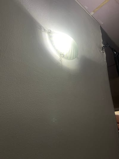 This media file depicts an indoor scene, likely a room with textured walls. The primary subject is a ceiling light fixture, which is illuminated and casts a bright glow, creating strong shadows on the wall. The light fixture appears to have a ribbed or grooved design. The walls are painted a light, textured color, possibly white or a light grey. In the upper left portion of the image, part of a ceiling or upper wall is visible, painted a muted purple color. A wooden beam is also partially visible along the edge of the purple surface. In the upper part of the image, a dark object, possibly part of a larger fixture or equipment, is visible, with some metallic elements and a grate-like structure.

The activity taking place is simply the illumination of the space by the light fixture. There are no people visible in the image. Notable details include the strong contrast between the bright light and the shadows, highlighting the texture of the walls. The lighting suggests it is likely nighttime or that the room has no other natural light source. There are no visible location cues beyond it being an indoor setting. The overall mood is neutral, with the focus on the lighting and texture of the environment. There is no visible text in the image.
