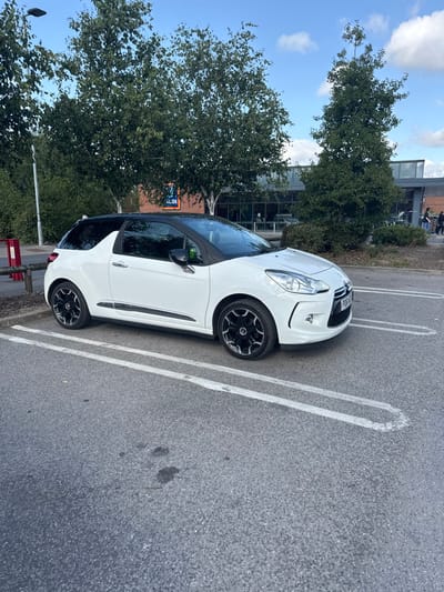 A white Citroën DS3 hatchback, UK registration PD61 NYF, is parked in an asphalt parking space marked with white lines. The vehicle features a black roof and black multi-spoke alloy wheels. In the background, the Aldi supermarket building at The Balk, Pocklington, United Kingdom, is visible, displaying its blue logo and a prominent glass facade. Deciduous trees with green foliage surround the supermarket building. Several indistinct human figures are discernible near the supermarket's entrance. The asphalt surface in the foreground exhibits two dark, irregular stains. A red and black bollard and a streetlamp pole are present on the left side of the frame. The scene is illuminated by natural daylight under a partially cloudy blue sky.