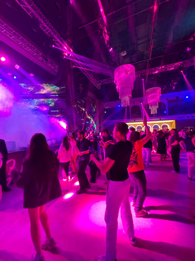 The image captures a vibrant indoor party or event taking place in Tsilkani, Georgia. The scene is bathed in intense pink and purple lighting, creating a dynamic and energetic atmosphere. A large crowd of people are visible, most of whom are dancing and enjoying themselves. In the foreground, a person in a black t-shirt and white pants is facing away from the camera, seemingly engrossed in the music. To their right, another person in an orange t-shirt and dark pants is also dancing. Multiple large, ornate chandeliers, illuminated with pink light, hang from the ceiling, adding a touch of grandeur to the venue. The background reveals more people, architectural elements like arches, and what appears to be a stage or backdrop with colorful light projections. The overall impression is one of a lively celebration with a focus on music and dancing.