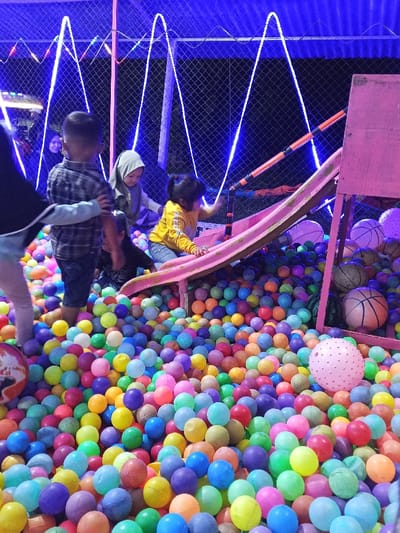 The image depicts a nighttime scene at a recreational ball pit located in Namplohbaro, Indonesia. The primary subject is a large enclosure filled with thousands of small, brightly colored plastic balls of various hues including pink, orange, yellow, green, blue, and purple.
Within the ball pit, several individuals are present. In the center-left foreground, a young boy, approximately 4-6 years old, wearing a plaid shirt and dark shorts, stands with his back to the camera. To his immediate left, a portion of an adult's body, wearing a dark top with white trim, is visible, with an arm appearing to interact with the boy. Further back and slightly to the right, another young child, possibly 5-7 years old, wearing a light-colored hijab, is partially submerged in the balls. Between these two children, a third, obscured individual in dark clothing is visible.
On the right side of the frame, a pink slide descends into the ball pit. A young girl, estimated to be 3-5 years old, wearing a yellow long-sleeved shirt and blue jeans, is positioned on the slide, facing towards the left, with her hands on the slide's edges. A pink, rectangular wooden-like structure stands adjacent to the slide on the far right.
The background features a chain-link fence, illuminated by prominent zigzagging blue LED strip lights and smaller multicolored fairy lights. Beyond the fence, several other individuals, including one wearing a dark head covering, are visible in the dimly lit distance.
Additional objects within the ball pit include several larger, spherical items near the slide structure on the right: two basketballs (one orange, one brown/tan), and another large, pale pink, spiky textured ball in the lower-right foreground. A red and white patterned ball is partially visible in the lower-left corner. The overall lighting suggests an artificial, nighttime environment.