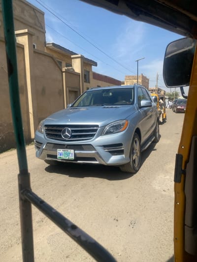A light blue Mercedes-Benz ML-Class SUV (W166 generation) is centered on an unpaved, dusty road in Sani Mai Nagge, Nigeria. The vehicle displays a Nigerian license plate, "ABUJA BWR-156CU," on its front bumper, which is equipped with a chrome lower trim. The scene is viewed from within a yellow tricycle taxi (keke napep), with a yellow frame and mirror on the right, and a green/grey metal bar on the left foreground.
The environment, under a clear blue sky, consists of single to two-story buildings with light brown or beige facades lining both sides of the street. Some structures show visible cracks in their walls. A modern street light pole is visible in the mid-ground. Behind the Mercedes-Benz, a second yellow tricycle taxi is present, followed by additional distant vehicles and multiple pedestrians. A faint silhouette of a driver is discernible inside the Mercedes-Benz. All vehicles appear to be stationary or moving slowly in a queue.