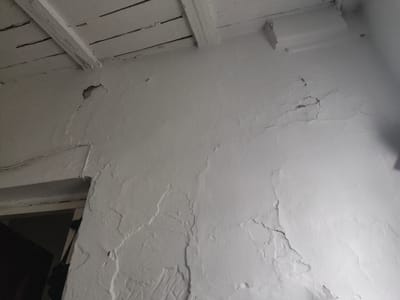 The image shows an interior view of a room with a damaged white-painted ceiling and wall. The ceiling is constructed of wooden beams and planks, some of which appear to be aged and possibly showing signs of wear or water damage. Cracks and peeling paint are visible on the wall surface, suggesting structural issues or moisture penetration. In the lower left corner, a doorway or opening leads to a dark interior space. The overall scene depicts a state of disrepair within a building, likely a residential or utility space. The location is specified as Ivanovka, Kyrgyzstan.