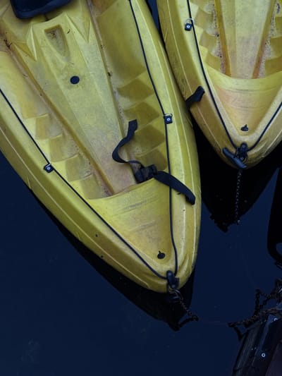 This is a close-up, overhead shot of two yellow kayaks floating in dark water. The kayaks are positioned next to each other, with the one on the left slightly closer to the viewer. Both kayaks appear to be made of plastic and have black ropes and straps attached to them. The water is calm and dark, reflecting the kayaks. The scene is devoid of human presence. There is no indication of a city or specific location within the image.
