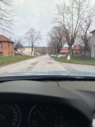 The image displays a forward-facing perspective from inside a vehicle, showing the dashboard, windshield wiper assembly, and partially visible speedometer and tachometer gauges in the lower frame. The scene is an unpopulated street in Suhache, Bulgaria, under an overcast sky. A worn, two-lane asphalt road extends into the distance, bordered by patches of green grass. Numerous bare deciduous trees line both sides of the road; those on the right have white-painted bases. On the left, a two-story brick building with a red tiled roof, appearing dilapidated with missing window panes, stands prominently. Further down this side, a yellow excavator is positioned near a lighter-colored building, and a blue car is visible on the road. On the right, a building with a light-colored facade and a red roof is discernible, with other residential structures further in the background. No people are visible.