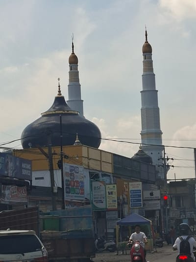 The image shows a street scene in Pantegajah, Indonesia, with a mosque prominently featured. The mosque has a large, dark dome and two tall minarets. The left minaret is white with gold accents, and the right minaret is also white with gold bands. In the foreground, there are several vehicles, including a truck, a car, and a motorcycle with a rider. A person on a red scooter is visible in the center of the frame, wearing a white shirt. To the right, another person on a motorcycle, wearing a white shirt and black backpack, is partially visible. Various signs and advertisements are displayed on buildings along the street, including one for "MATANG SERVICE" and another for "BSi MINI ATM". The sky is overcast with some clouds. The scene depicts a busy urban environment.