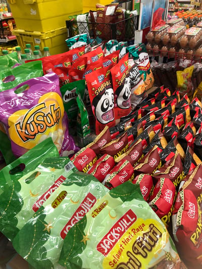 The image shows a close-up, eye-level view of shelves in a store, likely a minimarket or supermarket, in Putat, Indonesia. The shelves are filled with various packaged snacks, including chips and biscuits. In the foreground, several green and purple plastic bags with handles are visible, featuring prominent branding. To the right, stacks of red and dark red snack bags with yellow and white text are arranged in rows. Further back, there are yellow plastic crates and displays of drinks, as well as rows of bottled water. The overall environment is well-lit, suggesting an indoor retail setting. There are no people or active interactions depicted in the image.