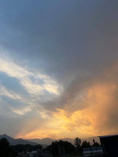 An outdoor, eye-level, wide shot captures a dramatic sunset over the city of Kabal, Pakistan. The sky is a canvas of vibrant colors, with streaks of orange and yellow clouds radiating from the horizon, transitioning to softer hues of blue and gray towards the top of the frame. Silhouetted mountains form a jagged line in the distance, their dark forms providing a stark contrast to the illuminated sky. Below the mountains, a line of dark green trees marks the edge of the city, with glimpses of buildings and structures peeking through. The foreground reveals the roof of a dark-colored building with a corrugated metal surface. The overall impression is one of a serene and breathtaking natural phenomenon unfolding over a quiet, rural landscape.