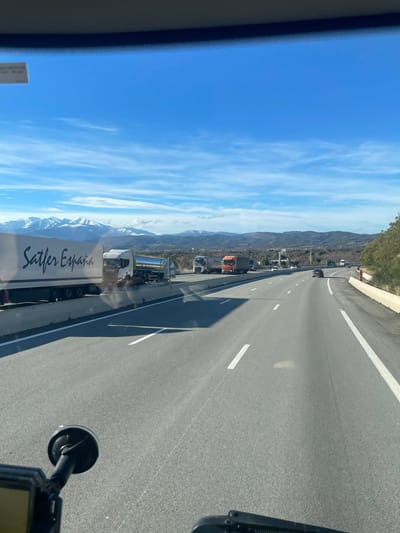 A highway scene shows multiple lanes, some occupied by trucks, including one labeled "Sattler España." Clear blue sky with scattered clouds covers the upper portion. Mountains are visible in the distance, partially obscured by haze. The view is taken from inside a vehicle. Road markings consist of white dashed lines. The road surface appears to be asphalt.