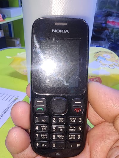 The image shows a close-up of a black Nokia mobile phone, held by a person's hand. The phone's screen is reflective and appears scratched. The keypad has numbers and letters in Cyrillic and Latin alphabets. The background is blurred, showing a green surface and a white object. A small pink sticker with "0%" is visible on the right side of the phone. The context of Ushtobe, Kazakhstan, is not visually represented in the image.
