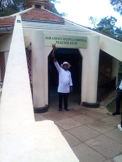 The image displays the Jaramogi Oginga Odinga Mausoleum, located in the South Sakwa ward of Bondo, Siaya County, Kenya. A male individual stands centered in front of the mausoleum's arched entrance, facing the viewer. He wears a light-colored long-sleeved tunic, dark trousers, dark shoes, and a light-colored brimmed hat. His right arm is extended upwards, holding a dark-colored, bushy whisk or fly-whisk. The mausoleum features light-colored walls, a prominent reddish-brown tiled roof with a small octagonal structure on top, and a sign above the entrance reading "JARAMOGI OGINGA ODINGA MAUSOLEUM." The ground directly in front of the entrance is paved with large square tiles. On the far right, partially obscured, are the lower legs and feet of a second individual wearing dark trousers and dark shoes. Green trees and foliage are visible in the background above the mausoleum.