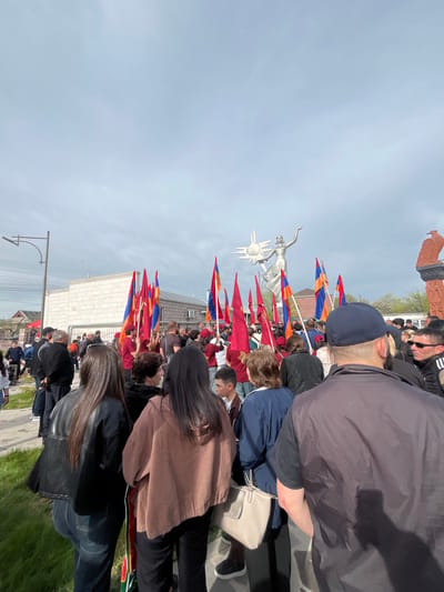 The image shows a daytime outdoor gathering of people in Akhuryan, Armenia. Many are holding Armenian flags (red, blue, and orange horizontal stripes). The statue "Mother Armenia" with a sun disk is visible in the background. The surrounding environment includes buildings, a street, and a grassy area. The photograph appears to have been taken from within the crowd.
