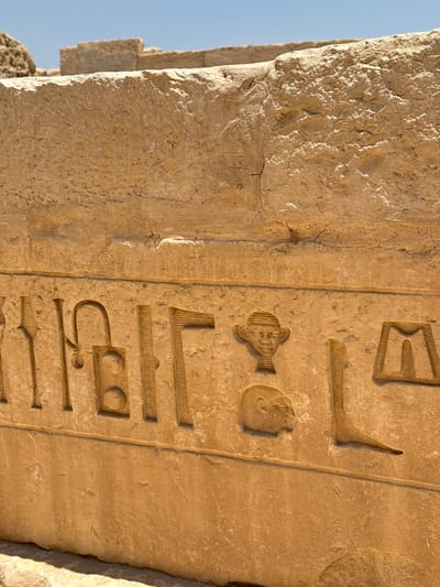 This is a close-up, eye-level shot of ancient Egyptian hieroglyphs carved into a stone wall. The hieroglyphs are weathered and appear to be part of a tomb or temple in Abu Sir, Egypt. The stone is a light tan color and shows signs of age and erosion. The hieroglyphs include various symbols, some of which are depictions of human heads and other figures. The background is a clear blue sky. The image focuses on the texture and detail of the carvings.