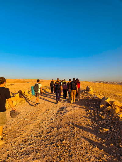 A group of approximately ten individuals are walking along a dirt path in a rocky, arid landscape under a clear blue sky. The sun is low, casting long shadows from the people and rocks. The individuals are wearing casual clothing suitable for hiking and several have backpacks, with one person carrying a rolled-up sleeping pad. In the distance, a city skyline is visible on a horizon line dotted with buildings. The terrain is characterized by dry earth, scattered rocks, and sparse vegetation. The scene suggests a hike or expedition in a desert or semi-arid region.
