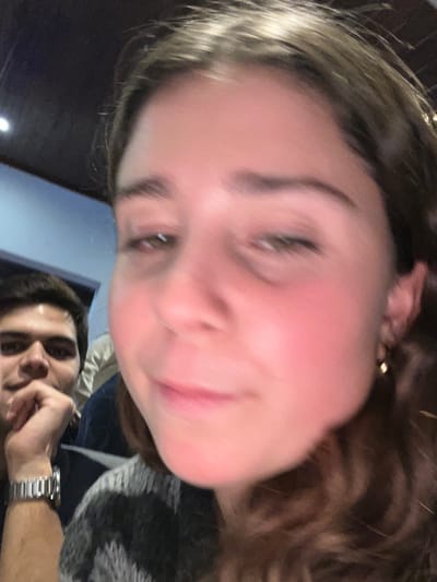 A selfie-style photograph shows a young woman with light brown hair and fair skin. She's smiling slightly. Visible behind and slightly below her is a young man with dark hair and a watch on his wrist. The background is dark. The image appears to be taken indoors.