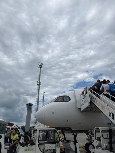 An overcast sky looms above an airport tarmac, where a white airplane is parked. Passengers are seen boarding the aircraft via mobile stairs. To the left, a control tower with a unique architectural design stands tall, resembling a bird or an owl. In the foreground, ground crew members are attending to airport equipment, including a tug vehicle. The scene captures a moment of departure or arrival at an airport, with the architecture of the control tower adding a distinctive touch to the environment. The presence of the plane, passengers, and ground crew indicates a busy aviation hub.
