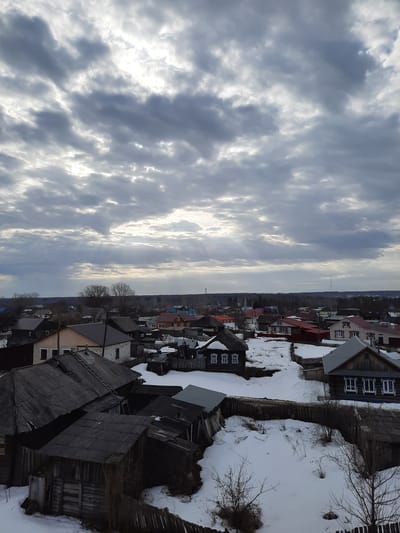 The image shows a winter scene in Kizner, Russia. The perspective is from an elevated position, possibly a hill or high building, overlooking a residential area. The foreground consists of a series of weathered, predominantly wooden houses with visible snow accumulation on the roofs and in the surrounding yards. The houses are closely packed, with varying roof styles and colors, although the overall color palette is muted due to the winter setting. Wooden fences and bare tree branches punctuate the landscape.

Beyond the immediate residential area, the view extends towards a distant treeline or forest, with a less distinct skyline. The sky occupies a significant portion of the frame, displaying a cloudy and somewhat overcast condition. The clouds are textured and diffuse, suggesting a soft, diffused light.