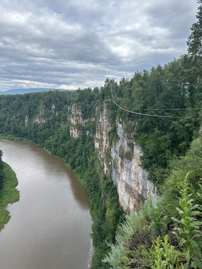 The image depicts a natural landscape with a dramatic cliff face overlooking a wide, calm river. The cliff is layered with white and brown rock, partially covered by lush green trees and vegetation that cascade down its slopes. A thin suspension bridge stretches across a section of the cliff, and a person wearing a bright pink garment is visible on it, suggesting an activity like ziplining or crossing the bridge. The river below is a murky brown color, reflecting the overcast sky. The sky itself is filled with textured, grey clouds, indicating a cloudy or overcast day. The overall scene conveys a sense of adventure and the grandeur of nature. The provided location context places this scene in Kul'metovo, Russia.