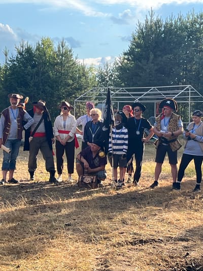 A group of eleven people dressed as pirates are standing and squatting outdoors in a grassy area with trees in the background. The sky is cloudy and blue. The pirates are wearing striped shirts, vests, bandanas, and hats. Some have eye patches and red sashes. One person is holding a pirate flag. There is a treasure chest in the foreground. The scene appears to be a reenactment or a themed event. The location is Дражнаўскі сельскі Савет, Belarus.