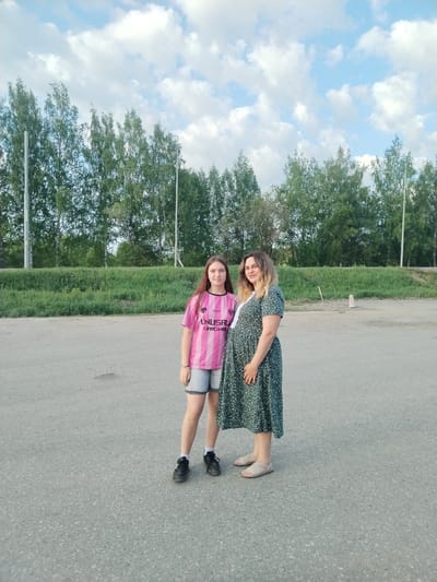 Two females stand on an asphalt surface under a partly cloudy sky, with a line of trees in the background. The younger female wears a pink striped shirt, denim shorts, and black shoes. The older female wears a green floral dress and sandals. The location is possibly Krilovo, Russia, based on the request. The asphalt surface appears to be a parking lot or road.