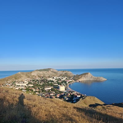 The image shows a panoramic view of the city of Ordzhonikidze, Ukraine, situated on the coast of the Black Sea. The scene is captured from a high vantage point, overlooking a settlement nestled between rolling hills and the expansive blue sea. The foreground is dominated by dry, golden grass, with the shadow of a person, likely the photographer, cast on the hillside.

In the midground, the town of Ordzhonikidze unfolds. Buildings of varying sizes and colors, predominantly white and red, are densely packed. Some structures appear to be residential homes with pitched roofs, while others are larger, multi-story buildings, possibly hotels or apartment complexes. Green trees and vegetation are interspersed throughout the urban landscape. A curved coastline is visible, with a small bay where boats are anchored. The water is a vibrant blue, reflecting the clear sky.

The background features rugged, light-colored hills that rise gently from the town and extend towards the sea. The terrain is characterized by its dry, arid appearance, with some evidence of erosion. The sky is a vast, cloudless expanse of bright blue, indicative of a sunny day. The overall impression is one of a coastal resort town bathed in warm sunlight.