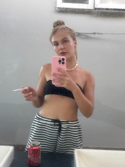 This media file is a vertical, full-body, mirrored selfie of a young Caucasian woman indoors. She is wearing a black bikini top, a white beaded necklace, and striped shorts. She has blonde hair pulled back and is holding a pink smartphone in her right hand, taking the picture. In her left hand, she holds a lit cigarette. Next to her, on what appears to be a counter, is a red can of what appears to be "DO" beer. The background is a plain, light gray wall, and there are some framed objects or windows on the left side of the image, which are reflective. The overall lighting suggests an indoor environment, possibly a bathroom or dressing room, with artificial lighting. The woman's expression is neutral. The location context provided is Van Hung, Vietnam, but there are no specific visual cues in the image to confirm this.